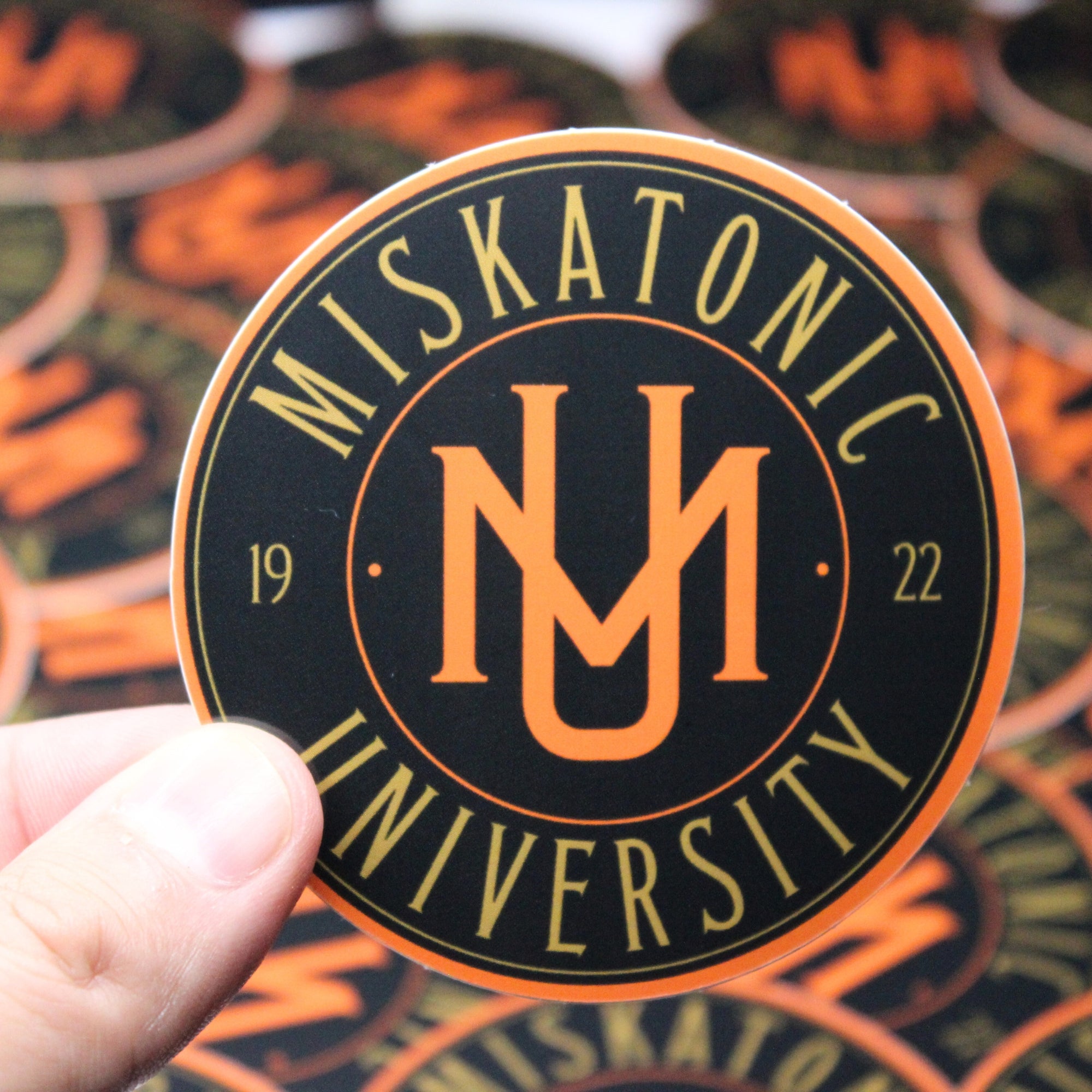 A hand holding a Miskatonic University coaster with an orange and black design.